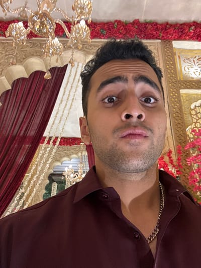 A young man is captured in a close-up, rotated portrait-style shot, looking directly into the camera with a distinct expression. He has a dark complexion, short, dark, wavy hair, and thick, dark eyebrows that are slightly raised, causing subtle wrinkles on his forehead. His brown eyes are wide, and his lips are slightly parted in what appears to be a surprised or contemplative pout. A light stubble is visible on his chin and jawline. He is wearing a dark maroon or burgundy collared shirt, and a shiny, golden-colored chain is visible around his neck.

The background suggests an ornate indoor setting, likely a venue for a formal event or celebration. Intricate golden architectural details, possibly carved wood or plasterwork with classical patterns, are visible behind him. Red floral garlands or arrangements run vertically along the left side of the frame and in the upper background. Below these, rich red drapes or fabric are visible. Decorative golden strings, possibly beads or crystal strands, hang in the mid-ground, contributing to a festive ambiance. A large, elaborate golden chandelier or light fixture, reflecting light, is partially visible in the lower-left corner. The overall lighting appears to be artificial, casting a warm glow on the scene. The man's pose and direct gaze indicate he is aware of being photographed, possibly taking a selfie.