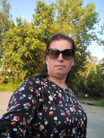 A woman is pictured in an outdoor setting, possibly a park or residential area, with lush green trees in the background. She is wearing a black floral print dress with red, white, and blue flowers and a gold chain necklace. She is also wearing large, dark-rimmed sunglasses. The sky is clear and blue, suggesting it is daytime. In the background, two large green trash bins are visible. The scene appears to be in the city of Kingisepp, Russia, given the context of previous images.