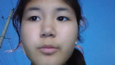 A close-up, head-and-shoulders selfie shows a young woman with dark hair and fair skin against a blue background. Her eyes are dark and looking directly at the camera. Her eyebrows are dark and arched. She has a small nose and full lips. She is wearing a small earring in her right ear. To the left of her head, a power line pole and wires are visible, suggesting an outdoor setting. The lighting suggests it is either dusk or dawn, or an artificially lit indoor environment. The blue background could be the sky or a wall.
