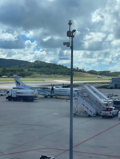 This image captures an airport scene in Mai Khao, Thailand, under a partly cloudy sky. A Bangkok Airways turboprop aircraft, identified by the registration "HS-PZK" and the airline's logo, is parked on the tarmac. A fuel truck, labeled "PTT-HKT-R02," is connected to the aircraft, indicating refueling operations are in progress. Mobile stairs, marked with "Vietjet Air" and various codes such as "TVI-PSD-005" and "TVI-PSD-008," are positioned next to the aircraft. A white pickup truck is parked near the stairs. In the background, lush green hills are visible, and another aircraft is seen on a distant runway. A tall pole with lights and a camera is prominently featured in the foreground, obscuring part of the sky and landscape. The overall environment is that of an active airport apron.