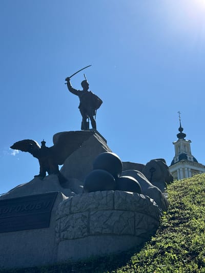 This is a low-angle, outdoor, full shot of a monument located in Vereya, Russia, on a clear, sunny day. The monument features a bronze statue of a soldier in military uniform, holding a sword raised above his head. The soldier stands atop a large rock formation. To the left of the soldier, a large bronze eagle with outstretched wings and a crown on its head is perched on a rock. In front of the eagle and the soldier, there are several large, dark, spherical objects resembling cannonballs. Below the main monument, a granite plaque with Russian inscriptions is visible. In the background, to the right of the monument, a white and yellow domed church with a cross on top is partially visible, rising above a grassy hill. The sky is a bright, cloudless blue, with the sun glinting in the upper right corner.