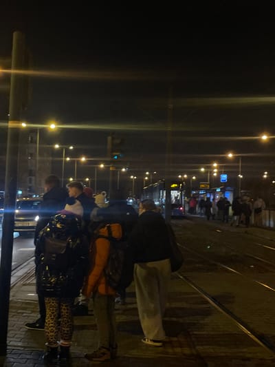 The image captures an urban public transport stop at night, likely a tram stop, illuminated by numerous artificial lights. Several people are gathered on a platform next to visible tram tracks, appearing to wait.
In the foreground, a group of individuals stands with their backs mostly to the viewer or in profile. One person wears a dark shiny jacket and distinctive leopard-print leggings. Another person in front of them has a bright orange jacket and a backpack. Further back, another person is seen wearing light grey pants and a dark top, also appearing to wait.
The scene is dominated by the dark night sky and the glowing streetlights and lampposts, which create prominent light flares. A blue rectangular sign, possibly indicating a stop or providing information, is visible in the mid-ground. What appears to be a tram or bus is partially visible behind the waiting crowd further down the platform. The overall setting suggests a bustling transit point in a city at night. No legible text is visible. The clothing worn by the people indicates cool or cold weather. This scene is consistent with a public transport location in a European city like Kraków, Poland.