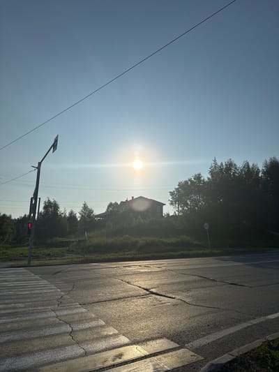 The image depicts a street scene in Koltsovo, Russia, at sunrise or sunset. A pedestrian crossing is visible in the foreground, with reflective white stripes on the cracked asphalt road. A traffic light on a pole shows a red pedestrian signal. In the background, a house with a dark roof is situated on a tree-lined hill. A speed limit sign indicating "40" is visible on the right side of the road. The sky is clear and bright, with the sun casting a lens flare. Power lines are strung across the sky.