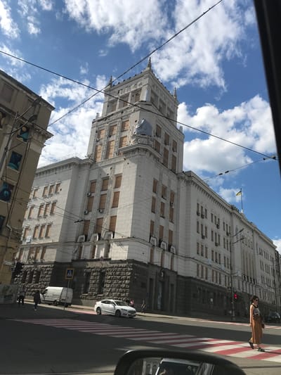 The image displays an urban street scene under a bright, partially cloudy sky. A large, ornate, light-colored building with classical architectural details dominates the left side of the frame. Many of its windows are boarded up with wooden panels. A yellow and blue flag, strongly suggesting the Ukrainian flag, flies from a pole on the building's facade near the top.
In the foreground and extending towards the right, a street features a prominent red and white striped pedestrian crossing. A red traffic light is visible, indicating that pedestrians should not cross. A person wearing a striped shirt and brown attire stands on the crossing, seemingly waiting. A white car and a white van are parked along the street. A blue and white pedestrian crossing sign is also visible.
Numerous black wires crisscross the sky, adding to the urban feel. A smaller, older building with some decorative patterns on its side is partially visible at the bottom. The photograph appears to be taken from inside a vehicle, as evidenced by a portion of a car's side mirror in the bottom right corner. The time of day is clearly daytime, with bright sunlight and shadows. The scene suggests a vibrant but possibly impacted urban environment, given the boarded-up windows.