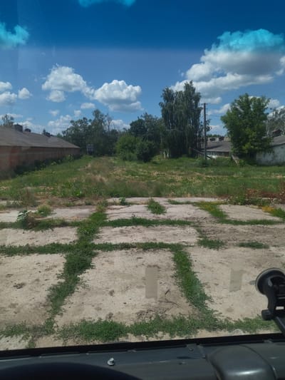 The image depicts an outdoor scene on a sunny day, likely in Buhakiv, Ukraine. The foreground is dominated by a cracked concrete surface with a geometric pattern of grass growing through the cracks, resembling a grid. In the middle ground, there is an expanse of overgrown green grass with scattered small yellow flowers. Behind the grass, there are several buildings with weathered roofs, some appearing to be industrial or storage structures made of brick and concrete. Tall trees, including a prominent birch tree, and other lush greenery are visible behind the buildings. The sky is a vibrant blue with fluffy white clouds, suggesting fair weather and midday. The photo appears to be taken from inside a vehicle, with the dashboard and a suction cup mount for a device visible at the bottom of the frame. No people are visible in the image. The overall impression is one of a somewhat neglected, rural or semi-industrial area bathed in bright sunlight.