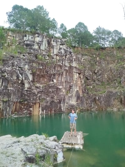 A man stands on a bamboo raft in a quarry lake in Lán Tranh, Vietnam. The man is wearing a blue t-shirt and khaki shorts, and he is holding a long pole. The raft is floating on the water, and the quarry walls rise steeply behind it. The walls are made of layered rock, and trees grow at the top of the quarry. The water is a deep green color. The sky is overcast.