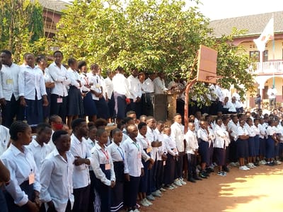 A large group of students, primarily female, are standing in rows on a dirt court in front of a building in Beni, Democratic Republic of Congo. The students are all wearing white shirts and dark skirts. There are several students in the background sitting on a low wall. A basketball hoop is mounted to a pole in the center of the court. The building behind the students appears to be a school with a flagpole and an African flag flying.