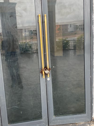 The image shows a close-up view of a double glass door, likely an entrance, with two vertical gold handles on each door. A rusty padlock with a chain secures the doors together, indicating they are locked. The glass is reflective, showing a distorted image of buildings, a body of water, and a railing on the other side. The floor inside appears tiled. The door frames are dark grey. No people are directly visible, but a shadowy reflection of a person in dark clothing can be seen on the left door. The setting is outdoor. The image is taken in Zanzi, Nigeria.