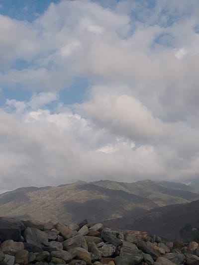 The image shows a rocky foreground with a mountainous landscape in the background. The sky is filled with a mix of blue sky and white clouds. The mountains appear to be covered in vegetation and are shrouded in some mist. The overall lighting suggests it is daytime. The scene is likely a natural landscape. It is not possible to determine the specific location in the city of Naiguatá, Venezuela based on the visual information provided.