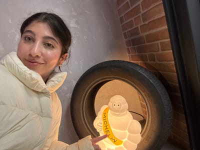 A female subject is prominently featured in an indoor setting in Alcobendas, Spain, positioned on the left side of the frame and smiling directly at the camera. She possesses dark hair, light eyes, and a philtrum piercing, and is attired in a cream-colored puffer jacket. Her right hand, with an extended index finger, is directed towards a white, illuminated Michelin Man (Bibendum) lamp. This lamp is situated within the circular opening of a dark vehicle tire, which rests against the background wall. The Michelin Man lamp displays "MICHELIN" on a yellow sash. The immediate background behind the subject and objects consists of a textured light grey wall on the left and a reddish-brown brick wall on the right, with a dark vertical element partially visible on the far right edge of the frame.