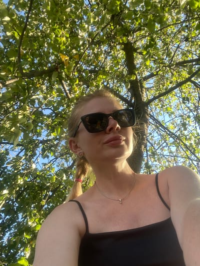 This is a low-angle, outdoor, waist-up, medium shot of a young woman, viewed from below, looking up. The scene is set in an apple tree grove under a bright, sunny sky, suggesting a warm, pleasant day. The woman is wearing sunglasses and a black top. Her blonde hair is styled in a braid. She has a delicate necklace around her neck. The tree is laden with small green apples and abundant foliage, indicating it is likely late spring or early summer. The camera angle creates a sense of looking up from the ground, perhaps as if the woman is lying down under the tree. The overall mood is relaxed and possibly playful. No text is visible in the image.