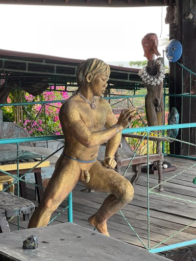 The image depicts an outdoor scene, possibly a balcony or terrace, in Tejakula, Indonesia. In the foreground, a golden-colored statue of a nude male figure in a running pose is prominent. The statue has its left leg forward, right leg back, and arms outstretched as if in mid-stride. A thin blue cord is wrapped around its waist. Behind the statue, a wooden post displays several decorative items. These include a wooden mask with what appears to be a long nose and red paint, a necklace of seashells, and a blue mask with wide eyes and painted features. Further back, there are glimpses of pink flowers, possibly bougainvillea, and lush greenery, suggesting a tropical environment. The ground is made of wooden planks. In the middle ground, a wooden chair with woven seating is visible, and to its right, a wooden carving of a human torso with shell adornments is displayed. To the far right, a dark, carved wooden object with a sculpted face is hanging from a wooden beam. The overall impression is of an outdoor space decorated with artistic and cultural artifacts.