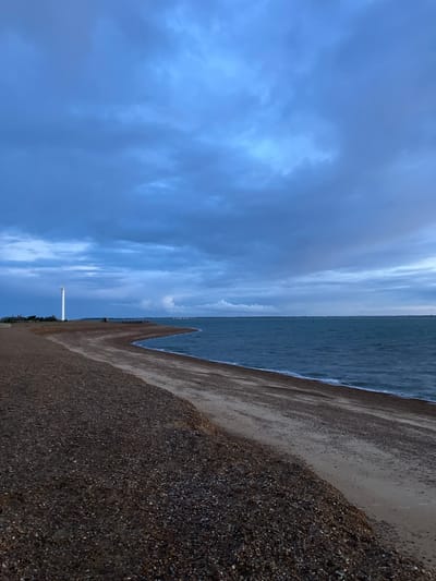 A serene coastal landscape is captured under a broad, overcast sky. The image portrays a tranquil scene featuring the sea meeting a pebble beach. The sky, occupying a significant portion of the upper frame, is filled with soft, textured clouds in various shades of blue and grey, indicating a cloudy or diffused light day.
A calm, dark blue-grey sea stretches horizontally across the midground, with gentle white-edged waves softly lapping at the shore. The foreground is dominated by a wide, textured pebble beach, transitioning from darker, coarser stones closer to the viewer to finer gravel and damp sand nearer the water's edge.
In the mid-distance on the beach, a tall, slender white pole or beacon stands vertically, a solitary man-made structure against the natural backdrop. Faint outlines of distant land or structures can be discerned along the far horizon line, where the sea meets the sky. No people or overt activities are visible, contributing to the peaceful and expansive atmosphere. The light is soft and even, consistent with an overcast day, without strong shadows to suggest a specific time of day. No visible text is present in the image. The setting evokes a sense of quiet solitude characteristic of a natural coastline, fitting the context of East Suffolk, United Kingdom.