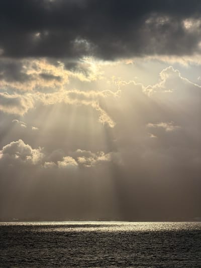 The image depicts a dramatic sky over a body of water. Dark, heavy clouds dominate the upper portion of the frame, with shafts of sunlight piercing through them, creating a crepuscular effect. These rays illuminate lighter, more scattered clouds below. The water in the foreground is dark and choppy, reflecting the light from the sky in a shimmering band along the horizon. A faint, hazy line of land with structures, possibly buildings, is visible in the distance along the horizon. The scene conveys a sense of natural power and atmospheric grandeur. The location is identified as Melaya, Indonesia, suggesting this is a coastal or offshore view near that area. No people or specific objects are discernible beyond the general elements of sky, water, and distant land.