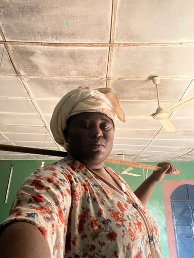 An adult female with dark skin, wearing a light-colored headwrap and a light-colored floral top with red, orange, and blue patterns, stands centrally in an interior room in Makera, Nigeria. She faces forward with a neutral expression, holding a light-brown wooden pole horizontally over her left shoulder with her left hand. Small hoop earrings are visible. The ceiling is composed of discolored square panels supported by visible battens, with a faint green circular mark on one panel. A cream-colored ceiling fan, inscribed "ESO OF C.S. YAKUBU SHADU" on a blade, hangs to the subject's right. The wall to the left is painted green. The wall to the right features a blue arched doorway or window frame with a light blue door or shutter. A dark brown horizontal beam is present in the mid-background.