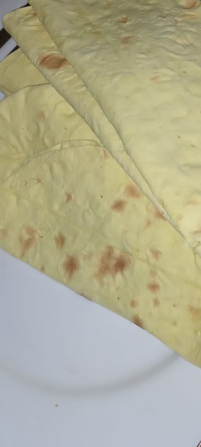 The image shows a close-up, overhead view of several folded flatbreads, likely lavash, stacked on a white plate. The flatbreads are pale yellow with irregular reddish-brown spots, indicating slight browning from baking. The texture appears soft and pliable. The plate is visible in the lower portion of the image and appears clean. There are no people or other objects in the frame. The scene is indoors, with neutral lighting that highlights the texture of the bread. There is no indication of the location being in Koviahy, Ukraine.