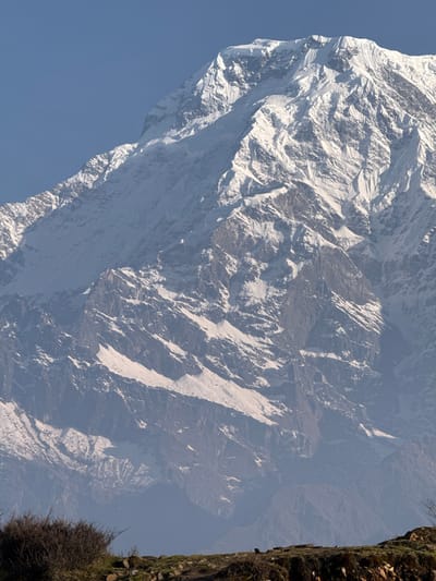 The image shows a snow-covered mountain peak with rock formations and a clear blue sky. The location is Lwang Ghalel, Nepal. The photograph is taken from a high vantage point, possibly from a cliff or another mountain peak. A rugged cliff with green moss and sparse vegetation is visible in the foreground to the right. The central focus is the colossal mountain face with snow and rock. The mountain's contours are sharp and well-defined, displaying a range of shades and textures created by the sunlight and shadow.
