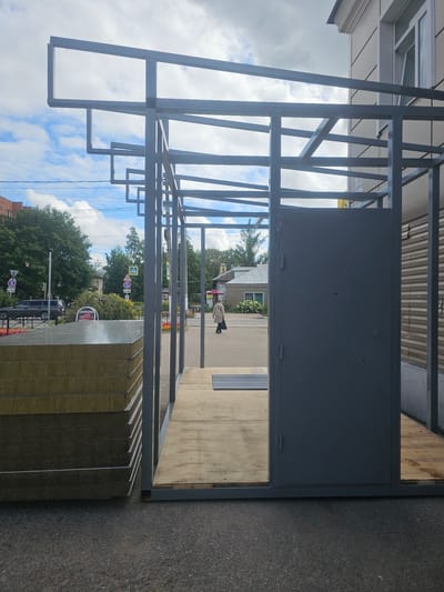An unfinished metal-framed structure with a gray door stands on a paved surface, possibly a sidewalk or street, in front of a building with a light-colored facade. Stacked construction materials, appearing to be insulated panels, are on the left side of the foreground. A person wearing a patterned coat and dark trousers is walking away from the camera in the middle ground. The background shows a street with trees, cars, and traffic signs under a cloudy sky. The overall scene suggests an urban construction site.