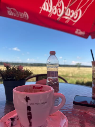 An outdoor scene in Lom, Bulgaria, features a reflective table under a bright blue sky with scattered clouds. In the immediate foreground, a white ceramic cup with a pinkish hue, marked "Buondi caffè" and "SAO PAULONDON" with a silhouette figure, sits on a matching saucer, containing beverage remnants. A small white sugar packet labeled "buondi" rests beside it. To the left, a small black pot holds an artificial plant with pink foliage. To the right of the cup, a dark-colored smartphone lies flat on the table. Further right, a plastic water bottle with a red cap stands partially filled, next to a glass with a dark straw, suggesting another beverage. The tabletop displays reflections and moisture droplets. In the midground, a blurry horizontal fence line separates the foreground from a vast, dry, yellowish-brown field with sparse vegetation under the clear sky. Overhead, a red umbrella prominently displays the white "Coca-Cola" logo multiple times. No individuals are visible.