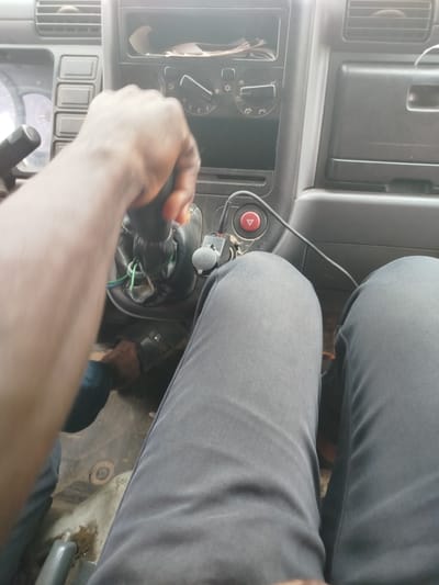 The image shows the interior of a vehicle, likely a truck, with a person's hands on the gear shift. The scene is viewed from the perspective of the driver or a passenger seated in the front.

The subjects are the hands and arms of a person, clad in dark clothing. The hands are actively engaged with the gear stick. The interior of the vehicle is mostly visible, featuring the dashboard with various controls, knobs, and vents. A red hazard light button is also visible. The seating area, covered by dark trousers, is also in view. The floor of the vehicle appears somewhat worn and dirty.

The activity taking place is likely driving or preparing to drive, as indicated by the grip on the gear shift. The focus is on the immediate controls of the vehicle.

Notable details include the textured dashboard, the design of the gear knob, and the presence of a 12V power outlet labeled "12V 60W". The lighting suggests it could be daytime, with ambient light filtering into the cabin. There are no strong indicators of the weather. The location context provided is Odiderengwaji, Nigeria, which does not offer specific visual cues within the image itself. No distinct emotions are conveyed.