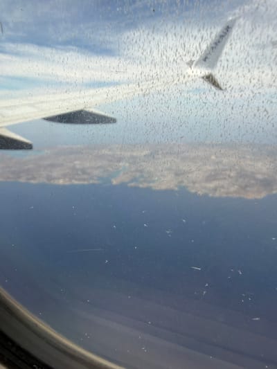 The view from an airplane window shows a Ryanair wing in flight. The window is dirty, with many small smudges and scratches. The sky and clouds are visible through the window. The location is inferred to be Luqa, Malta, based on Ryanair's extensive operations at Malta International Airport.