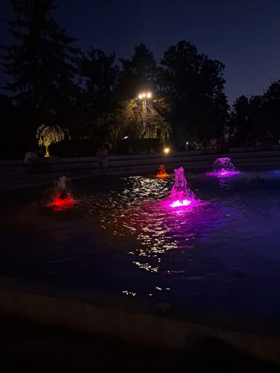 A night scene at a fountain in the city of Popovo, Bulgaria, features illuminated water jets in vibrant colors. The fountain's water is lit with red, pink, and purple lights, creating a dramatic effect against the dark water. In the background, silhouetted trees and a lamppost with warm yellow light are visible. Several people are seated and standing on a tiered stone structure behind the fountain, appearing to observe the display. The sky above is a deep, dark blue, indicating it is nighttime. The foreground shows the edge of the fountain's basin and the dark ground surrounding it.