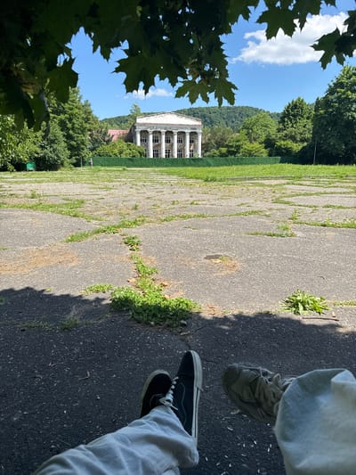This is a vertically oriented, full-shot photograph taken outdoors on a sunny day. In the foreground, the legs and feet of two people are visible, one wearing light-colored pants and grey athletic shoes, the other wearing grey pants and black canvas sneakers. They are standing on a cracked asphalt surface with patches of grass growing through the cracks.

The middle ground features a large, white, neoclassical building with a colonnade of tall pillars. The building appears to be in a state of disrepair, with some signs of weathering. In front of the building is a wide expanse of green grass, and beyond that, a dense forest covers a hillside. The sky is a clear blue with a few fluffy white clouds.

In the far left foreground, the dark green leaves of a tree partially obscure the view. The overall scene suggests a park or a historical site. The lighting indicates it is daytime, likely late morning or early afternoon, with bright sunlight casting shadows. The weather appears to be warm and pleasant. The presence of the building and the surrounding nature points to a location with historical significance. No text is visible in the image.