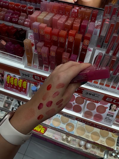 The image shows a close-up of a person's arm with several lipstick or lip gloss swatches applied in a store in Nong Pa Khrang, Thailand. The arm is positioned in front of shelves stocked with various makeup products, predominantly lipsticks in clear tubes with colorful contents. Many of the lipsticks have labels indicating brand names and shades, such as "Jelly Tint," "Try Me," and numerical designations. There are also displays of blushes and compact powders. The lighting suggests an indoor retail environment. The swatches on the arm range in color from light peachy tones to deep reds and berries.