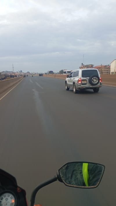 A silver SUV drives on a multi-lane road under a cloudy sky. The road surface appears wet, with a faint streak running down the center lane. In the distance, a gathering of people stands by the roadside. In the foreground, the motorcycle's speedometer and side mirror are visible, the mirror reflecting a bright green object. Buildings can be seen on the side of the road.
