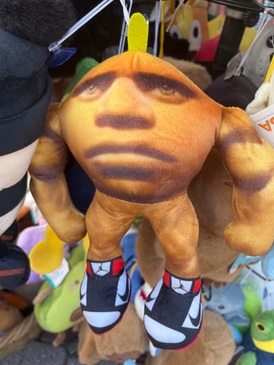 The image shows a close-up of a tan-colored plush toy with a human-like face and dark brown stripes on its head and limbs. It has a green stem-like protrusion on its head and is wearing black and white sneakers with red accents and a black Jumpman logo, resembling Air Jordan 1s. The toy is hanging among other plush toys in what appears to be an outdoor market stall, possibly in Potos, Greece. The background is blurred and filled with various other plush toys, some of which are brightly colored. The lighting suggests it is daytime.