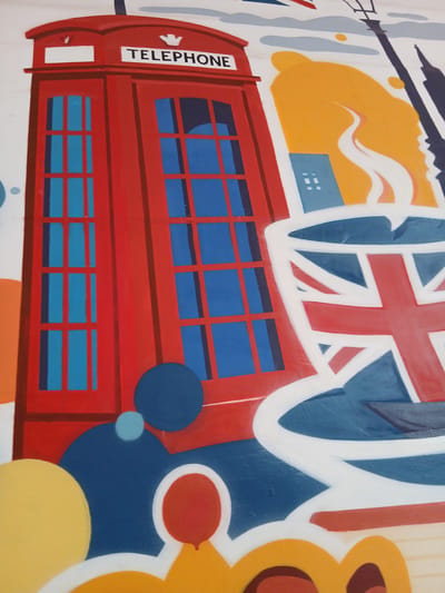The image displays a vibrant, pop art-style mural or graffiti on a wall, likely located in Dupnitsa, Bulgaria, although its subjects strongly evoke cultural symbolism of the United Kingdom. The central and most prominent subject is a classic red British telephone booth, depicted in a simplified, graphic manner with bright blue windows. The word "TELEPHONE" is written vertically in white letters on a rectangular panel on its left side.
Above and partially behind the telephone booth, a large Union Jack flag is rendered in bold white, red, and blue, creating a dynamic backdrop. The background features a mix of abstract shapes and colors. To the upper left, a dark silhouette, possibly representing a famous London landmark like Big Ben or a similar tower, stands against an irregular, yellow and orange cloud-like shape. White, wavy forms, suggestive of clouds or steam, are interwoven with the colorful elements, creating a sense of movement. Smaller blue rectangular shapes, reminiscent of building windows, are also present in this section.
The composition includes various circular forms in different shades of blue, orange, and yellow, adding to the dynamic and playful aesthetic. On the far right, a large, stylized yellow and brown element hints at a cartoon-like figure or abstract sun. The mural's style is characterized by strong outlines, flat color blocks, and a lively palette, creating a strong visual impact. No human figures are present, and the scene is static, capturing a moment of artistic expression. The time of day and weather are not explicitly conveyed, given the abstract nature of the background elements.