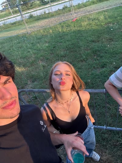 The media file is a selfie taken outdoors, likely in Segrate, Italy. Two people are visible: a young woman in the foreground and a young man's face in the bottom right corner. The woman has blonde hair and is wearing a black top, blue jeans, and a necklace. She is puckering her lips for the camera. The man has dark hair and is partially visible, looking towards the camera. They appear to be at an outdoor event or gathering, possibly a festival or concert, as indicated by the presence of a metal barrier and a grassy area that looks like a park or field. The lighting suggests it is late afternoon or early evening, with warm sunlight casting a glow. The woman is holding a translucent cup with a green liquid, and the man is holding a cup with a red liquid. The overall mood seems relaxed and casual. There is no visible text in the media file.