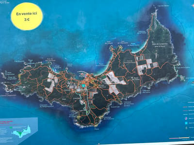 The image is a detailed map of Port-Cros island, located in Hyères, France. The map is a signpost, likely placed outdoors for tourists. It depicts the island's topography, with various geographical features labeled in French, such as capes, beaches, forts, calanques, and hamlets. There are also marked hiking trails of different colors, indicating their type or difficulty. A prominent yellow circle on the upper left corner of the map states "En vente ici 3 €" (On sale here 3 €), suggesting that tickets or guides are available for purchase at this location. A small inset map in the bottom left corner shows the location of the islands of Port-Cros and Porquerolles within the Var region. The map includes a legend with symbols for trails, a tourist office, and a house, as well as a scale bar. The overall environment is a marine landscape with clear blue waters surrounding the island. The map appears to be made of a durable material suitable for outdoor display, and there is a protective plastic film over the yellow "En vente ici" circle.