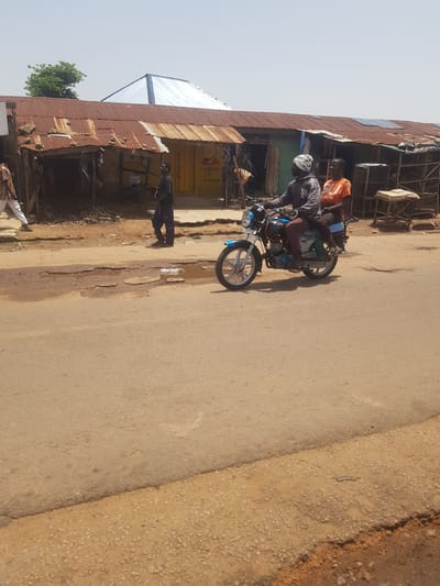 In Tukur Tukur, Nigeria, a motorcycle with two occupants travels on a paved road. The driver, a man wearing a dark patterned shirt and a white headscarf, operates the motorcycle. Behind him, a passenger, a woman wearing an orange t-shirt, sits on the back. The road is in poor condition, with several large potholes filled with murky water. In the background, a row of buildings with corrugated iron roofs and weathered walls lines the street. Some of the buildings appear to be shops, with open fronts displaying various goods or services. A man in a striped shirt and white pants walks on the far left of the image, and another man in dark clothing walks on the right. The sky is clear and the sun is bright.