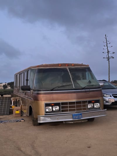 A wide shot captures a vintage, two-tone brown and gold RV parked on a dirt lot under a cloudy, overcast sky. The RV, with its distinctive chrome grille and large panoramic windshield, dominates the foreground. To its left, several solar panels are propped up. In the background, a silver sedan with a license plate reading "8NEZ253" is parked to the right of the RV. Further behind, a tall, bare agave plant stands silhouetted against the sky, and a distant building with trees is visible. The license plate on the RV reads "1KVX720". The overall scene suggests an outdoor, possibly rural or undeveloped area, consistent with a location on the outskirts of San Diego, California, United States of America, perhaps an RV park or a private property where alternative living arrangements are possible. There are no people visible in the image.