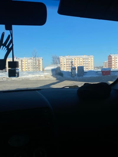 The image displays a daytime winter scene in the city of Dokshytsy, Belarus, viewed from inside a vehicle. The upper portion of a dark dashboard occupies the lower half of the frame. A dark rearview mirror hangs centrally from the top, with a black air freshener shaped like two hands and a small black rectangular bottle suspended below it. A dark, soft object, possibly a hat, rests on the right side of the dashboard.
Outside the vehicle, the foreground features a paved area, partially clear of snow, bordered by significant snowdrifts. Two snow-covered structures resembling fuel pumps or industrial containers are visible on the right, along with a smaller, red, snow-covered cylindrical object, possibly a waste bin. A large, snow-covered, arched structure is present on the left.
The background is dominated by a clear, vibrant blue sky. Several multi-story residential buildings are prominent: a six-story light yellow apartment building is on the left, and a larger seven-story light yellow apartment building with a snow-covered roof is in the center-right. A portion of a brown/red multi-story building is visible on the far right. Bare deciduous trees are interspersed within the snow-covered landscape between the structures. The scene is static, with no individuals or active vehicles visible.