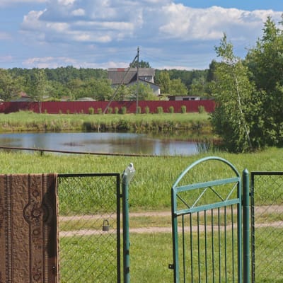 The image captures a rural scene in Блужскі сельскі Савет, Belarus. In the foreground, a dark, intricately patterned rug is hung on a chain-link fence, partially obscuring the view. To the right, a green metal gate stands open, revealing more of the fence and the grassy area behind it. A small padlock is attached to the fence.
Beyond the fence lies a body of water, likely a pond or small lake, bordered by lush green grass. In the middle ground, a red metal fence runs horizontally, separating the grassy area from the background. Behind the red fence, several buildings are visible, including a gray structure with a distinctive red roof and a power line pole. The landscape is dotted with trees, and a bright blue sky with scattered white clouds completes the scene. The overall impression is one of a tranquil, undeveloped rural environment.