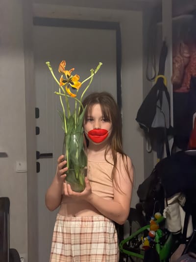A young girl stands indoors, holding a vase of wilting orange tulips. The vase is glass and cylindrical. The girl wears a beige top and plaid bottoms. Over her mouth, she has a pair of red, oversized lips. Behind her is a shelf with items, including a black bag, a stroller, and other indistinct objects. The lighting is dim. The location is Tokkari, Russia.