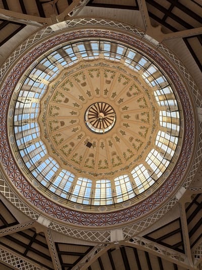 The image shows a detailed view of the interior dome of the Central Market in Alboraia, Spain. The dome features intricate ornamentation, including painted floral motifs of branches with yellow fruit and green leaves. The dome is constructed with a series of segmented windows that allow natural light to enter, creating a bright and airy atmosphere. The windows are arranged in a circular pattern around the apex of the dome. The structure of the dome is supported by visible beams and metalwork, forming a complex architectural design. The overall impression is one of grand and ornate architecture, likely a historical building.