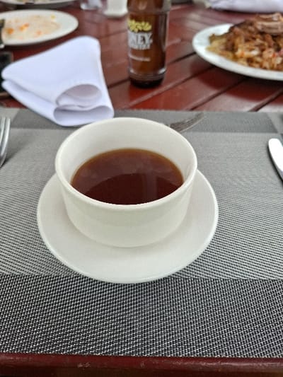 A white cup and saucer containing dark red liquid, presumably tea, are situated on a white and grey woven placemat. The placemat rests on a brown wooden table, likely in an outdoor dining area. A partially visible bottle of brown liquid and a white napkin are present on the table. The scene is most likely a restaurant or cafe in Karagita, Kenya, a suburb of Nairobi. The coordinates (-0.773, 36.42) suggest a location close to the Karagita Ridge, which is known for its scenic views and numerous restaurants.