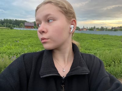 A young Caucasian female with blonde hair pulled back is pictured outdoors. She is wearing a black fleece jacket, a silver necklace with a single pendant, and a white wireless earbud in her right ear. She is looking to her left, away from the camera. The background consists of a green field with some purple flowers, a light blue fence, trees, and a few houses. The sky is cloudy with a hint of sunset colors. The location is specified as Klepiki, Russia.