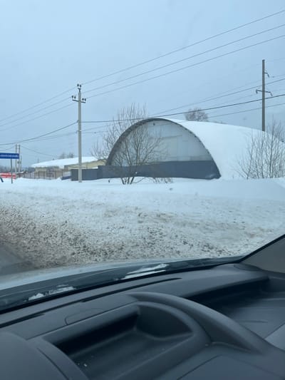 The image captures a snow-covered winter scene in Poddubki, Russia, viewed from inside a vehicle. The lower frame reveals a dark gray vehicle dashboard and the base of the windshield, with minimal ice or snow visible along its edge. Directly outside, a substantial mound of dirty, brown-tinted snow occupies the foreground, suggesting roadside accumulation or slush.
Beyond this snow, a dark gray corrugated metal fence stretches across the mid-ground. Several bare deciduous trees and shrubs are scattered in front of and behind the fence, some partially covered by snow. The most prominent structure is a large, arch-roofed building, typical of a quonset hut, positioned to the right of the frame's center. This building features white or light gray corrugated metal paneling on its upper sections and a darker gray base, with its entire curved roof heavily blanketed in a smooth layer of white snow.
The background consists of an overcast, light gray sky. Multiple horizontal power lines are visible, supported by two concrete utility poles. To the far left, a blue road sign with white Cyrillic text, partially decipherable as "Г-ВНУКОВСКИЙ" (G-Vnukovsky), is visible above the snow line, with a red sign beneath it. Another white sign with black Cyrillic text, possibly indicating "БРИКЕТЫ" (Briquettes) and "КУПИТЬ" (Buy), is positioned behind the fence line, to the left of the arched building. No individuals, animals, or discernible actions are present in the scene. The environment is consistent with a wintry, semi-rural or industrial roadside setting.