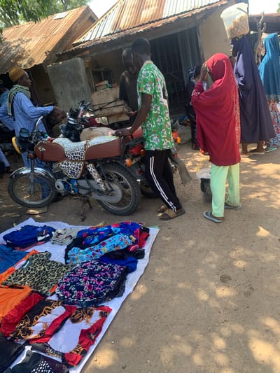The image depicts an outdoor scene, likely a market, in Boi A, Nigeria. The ground is dirt, with sunlight casting dappled shadows, suggesting it is daytime. Several people are visible, mostly dressed in traditional African attire. One person, wearing a red garment and light green pants, appears to be attending to something on a motorcycle. Another individual in a blue shirt and yellow head covering is seated. The focal point includes several motorcycles and a spread of colorful clothing, including patterned dresses and scarves, laid out on a white cloth, implying a commercial activity or sale. The setting includes a corrugated metal structure on the left, possibly a shop or dwelling. The overall mood appears to be bustling and active, characteristic of a market environment. There is no discernible text in the image.