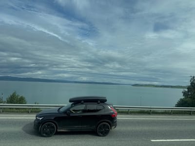 A black Volvo XC40 SUV with a black cargo box on its roof is driving on a paved road in Skogn, Norway. The car is black and has black wheels. The road has white lane markings. A metal guardrail is visible to the left of the road, with grass and trees behind it. Beyond the guardrail, a large body of water, likely a fjord, stretches to the horizon. Hills and distant land are visible across the water. The sky is cloudy and overcast. The image appears to have been taken from another vehicle moving in the same direction.