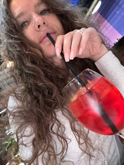 A close-up, indoor, selfie-style shot shows a woman with long, curly, brown hair holding a glass of red liquid with a black straw. She is wearing a light gray sweater and a ring with two bands on her right ring finger. Her lips are slightly parted, and she is looking towards the camera. The background is blurred, but suggests an indoor setting with some lighting and possibly a screen in the upper right corner. The image appears to have been taken in the city of Dubrovka (Lunovskoe S/P), Russia.