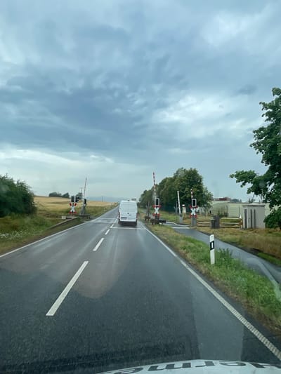 A white van drives towards a railway crossing on a wet asphalt road under a cloudy sky. The van is in the right lane of a two-lane road, which has white dashed lines marking the lanes. On the left side of the road, there is a field of yellow crops. On the right side of the road, there is a sidewalk, grass, and trees. The railway crossing has red and white striped barriers, red and white striped poles, and flashing lights. There are also buildings in the background on the right side. The scene appears to be in a rural area, possibly on the outskirts of Lauterbach, Germany.