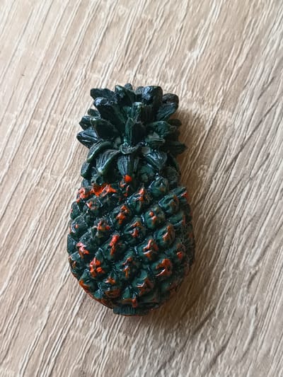A small, dark green, textured object resembling a pineapple is positioned centrally on a light brown wooden surface. The object has red accents on its surface, highlighting its hexagonal patterns. The wooden surface displays a prominent wood grain pattern with diagonal lines. The image is a close-up shot, focusing solely on the pineapple-like object and the surface it rests upon. There are no people or any other objects visible in the frame. The scene is static and appears to be an indoor setting. There is no information in the image to suggest a location in Tebras, Latvia.