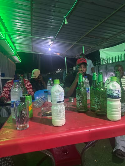 A medium shot depicts a red plastic table laden with various beverage containers, indicative of a social gathering. Dominating the tabletop are several green glass bottles of Heineken beer, alongside plastic bottles filled with water and a white, opaque liquid labeled "Fresh Yoghurt." Small metal caps and miniature glasses are scattered nearby, suggesting recent consumption.
Several individuals are partially visible around the table. One person is seen wearing a red cap, while another dons a white baseball cap with the letter "G." The attire of others includes patterned sweaters and casual shirts. The setting appears to be outdoors, possibly at night, under artificial lighting, judging by the illuminated metal framework and green-lit wall in the background. While the exact location remains ambiguous, the "Queen's Land Fresh Yoghurt" bottle suggests the setting is in Nigeria. The city of Kurudu is provided contextually.