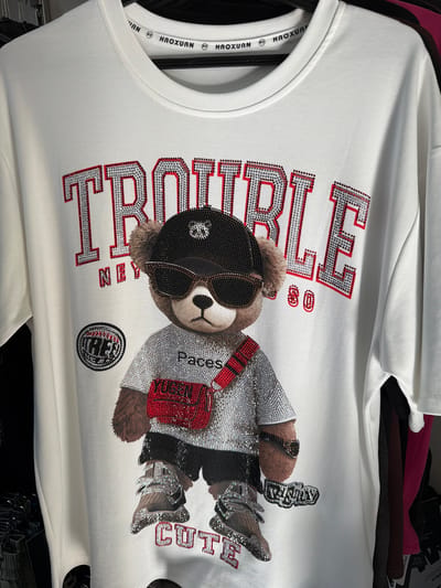 This media file shows a close-up, slightly angled shot of a white t-shirt with a large graphic design. The graphic features a cartoon bear character embellished with rhinestones. The bear is wearing sunglasses and a hat, both also decorated with rhinestones. It is holding a red bag and has its arms around itself. The t-shirt has text printed on it in large, rhinestone-covered letters. Some of the text visible includes "TROUBLE," "AIN," "Paces," and "CUTE." The t-shirt also has a brand tag on the inside of the collar with the text "HAOXUAN" repeated. The overall style of the graphic is bold and flashy, with a playful and somewhat edgy aesthetic. The t-shirt appears to be new, possibly displayed for sale. There are no other people or a discernible outdoor scene. The lighting is bright, suggesting an indoor setting, likely a retail environment.