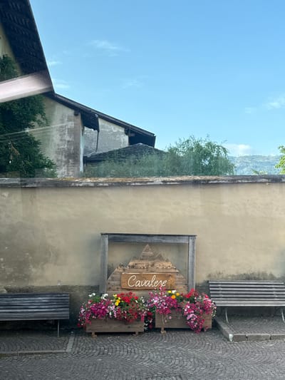 A low-angle, outdoor, full shot captures a town square in Cavalese, Italy, under a clear blue sky with sparse clouds. In the foreground, a cobblestone path leads to two weathered, wooden benches positioned on either side of a central wooden planter box. The planter box, adorned with colorful pink, red, and yellow flowers, sits within a rustic wooden frame. A wooden sign with the name "Cavalese" and carved mountain imagery is mounted on the frame above the flowers. Behind the planter and benches, a pale yellow, textured stucco wall stretches across the mid-ground. Above the wall, the upper portions of old, stone buildings with dark tile roofs are visible, partially obscured by lush green trees. In the distance, hazy, grey mountains rise against the sky. The scene is devoid of any people or visible actions.